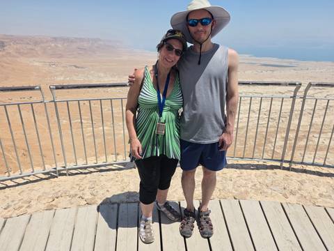       Two tourists stand arm in arm on a wooden platform overlooking the sun-baked desert landscape and Dead Sea basin.
  