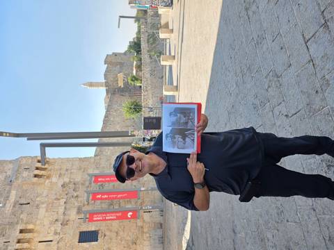       Tour guide smiling while holding historical photos in front of ancient stone walls and a tower in Jerusalem.
  