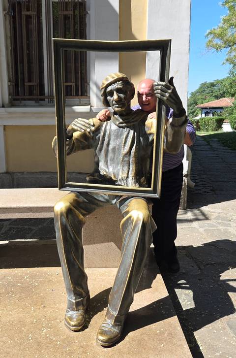       Playful bronze statue of a seated man holding an empty picture frame, visitor’s face appearing through the frame.
  