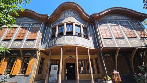       Beautifully painted Bulgarian Revival house with curved wooden eaves and decorative motifs in a leafy courtyard.
  