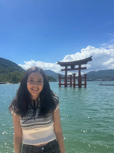       Smiling traveller with the iconic red torii gate of Itsukushima Shrine rising from the sea behind her.
  
