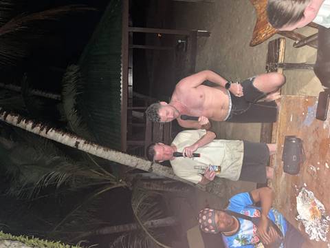       Two men singing karaoke with microphones on a tropical beach at night, illuminated by camera flash.
  