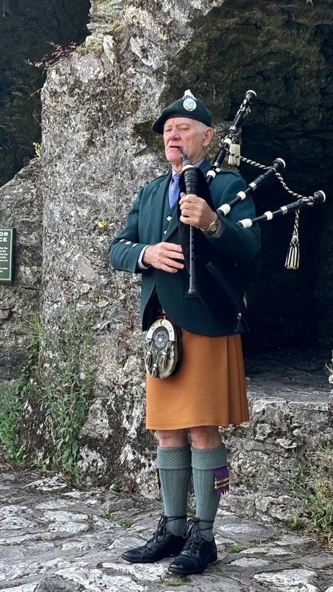       Traditional piper in kilt and jacket playing bagpipes beside ancient stone walls.
  
