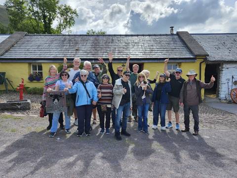       Large tour group cheering outside a quaint yellow-walled thatched cottage museum.
  
