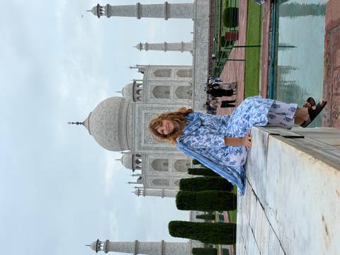       Woman in floral attire poses at marble bench with Taj Mahal rising majestically behind.
  