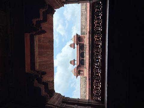       View of a red-sandstone pavilion framed by an ornate arch with blue sky and clouds in the background
  
