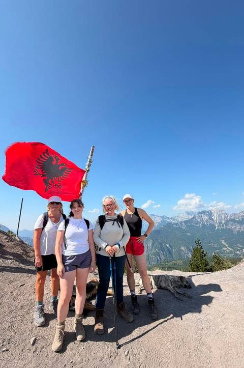       Four hikers posing at a mountain summit beside a large Albanian flag with alpine peaks behind.
  