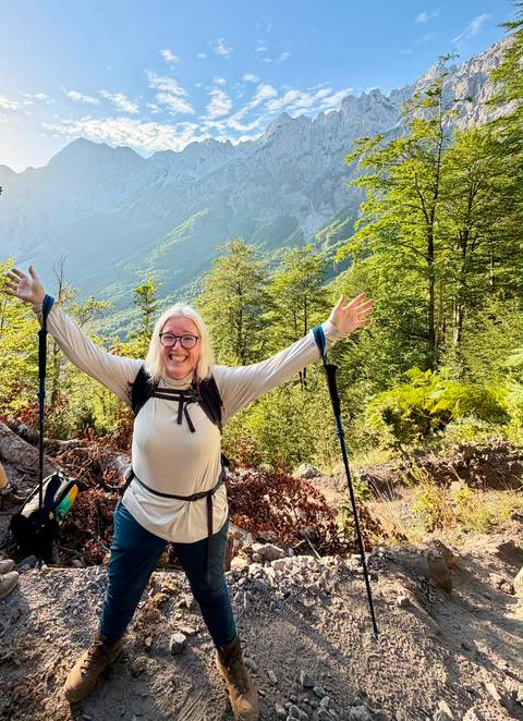       Cheerful hiker with trekking poles raises arms in a lush alpine forest with mountains beyond.
  