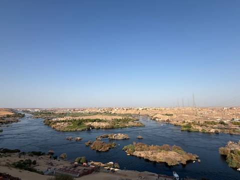       Wide view over islands and blue waters of the Nile with a distant desert town under clear sky.
  