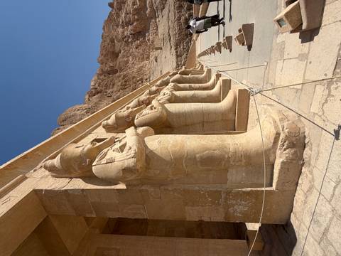       Row of sandstone statues with crossed arms guarding the terrace of an ancient mortuary temple against desert cliffs.
  