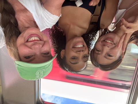       Three women smiling for a selfie inside a moving train in Japan, with a red train blurred outside the window.
  