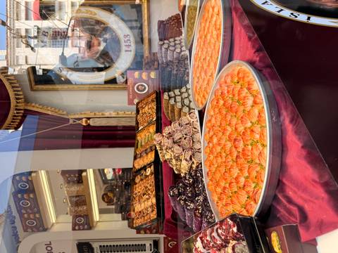       Large trays of bright orange baklava and shelves of Turkish sweets displayed in a shop window.
  