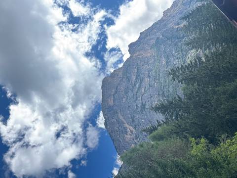       Towering rocky mountain peak framed by evergreen trees and scattered clouds.
  
