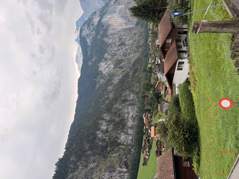       Alpine village houses on grassy slope beneath high cliffs and overcast sky.
  