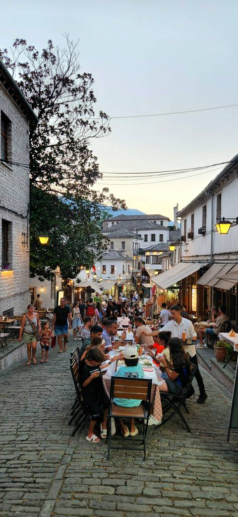       Bustling evening bazaar street lined with white stone buildings, lit shopfronts and crowds of visitors.
  