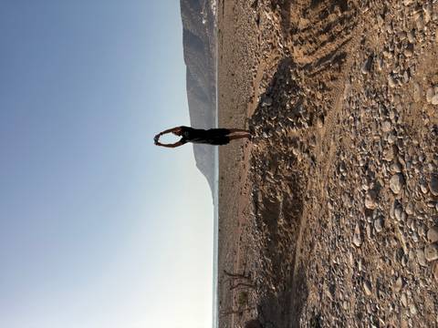       Person standing on stony ground stretching arms toward the sky beside a rugged coastline.
  