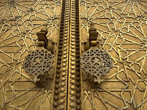       Ornate golden doors of a Moroccan palace with intricate metalwork knockers and geometric patterns.
  