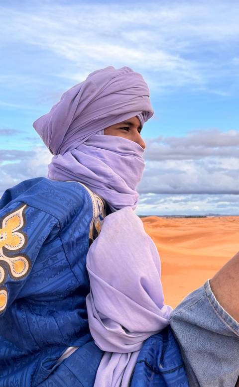       Side profile of turban-clad traveller smiling against sweeping Sahara sandscape.
  