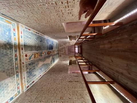       Long wooden walkway inside an Egyptian tomb with colorful hieroglyphic ceiling and illuminated walls.
  