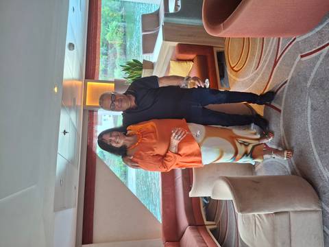       Smiling couple holding wine glasses in a river-cruise lounge with water visible through windows.
  
