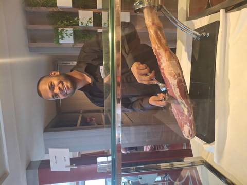       Chef slices cured ham behind a glass counter, smiling at the camera.
  
