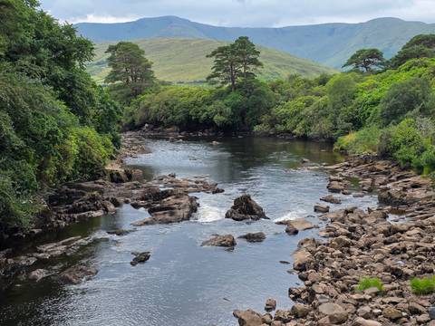       Serene river winding through rocky shallows with lush green hills in the distance.
  