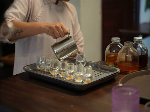       Hand holding a metal pitcher pouring amber coffee into miniature glasses beside syrup bottles.
  