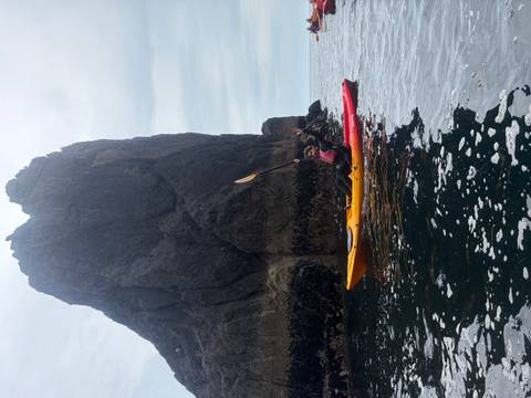      Kayaker paddles beside a dark sea stack, raising a triumphant hand on the Atlantic.
  