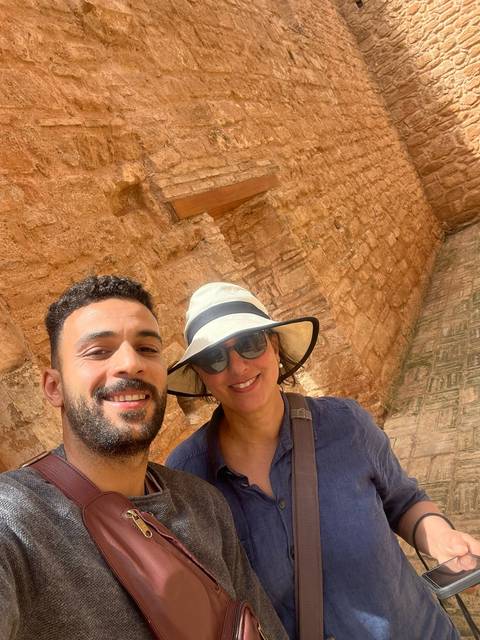       Smiling travellers take a sunny selfie in front of ancient sandstone walls with ornate carvings.
  