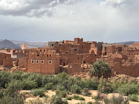       Panoramic view of the adobe ksar of Aït Benhaddou with palm groves in the foreground and arid hills beyond.
  