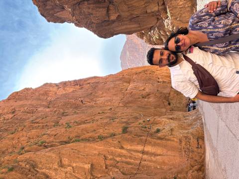       A smiling couple stand beside the towering orange cliffs of Todra Gorge under a bright sky.
  