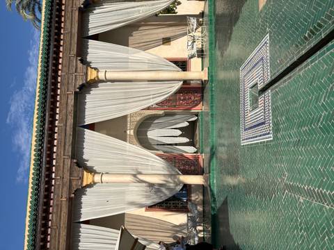       Elegant riad courtyard with green-tiled pool, cream drapes and carved columns highlighted by afternoon sun.
  