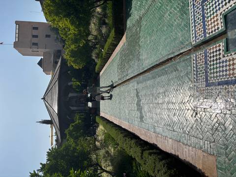       Long tiled pathway bordered by lush gardens leading to a pavilion where two visitors stroll in the sun.
  