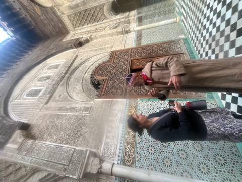       Blurred image of a guide explaining ornate madrasa architecture to a visitor within a tiled courtyard.
  