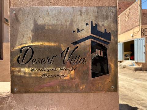       Weathered brass sign for 'Desert Villa Boutique Hotel Merzouga' mounted on a wall in desert light.
  