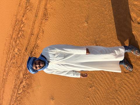      Berber guide in white djellaba and blue turban standing on golden sand with dune tracks behind.
  