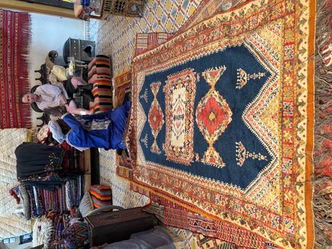       Local vendor displays a large patterned carpet while a traveller examines it inside a colourful showroom.
  