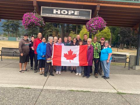       Smiling tour group posing with a large Canadian flag under a wooden shelter adorned with hanging flower baskets.
  