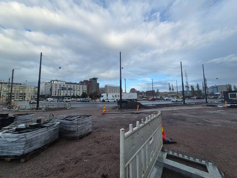       Construction site with scattered pallets, machinery and city buildings under a grey sky.
  