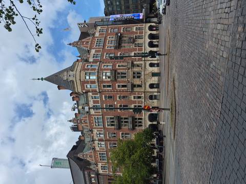       Grand red-brick historic building with ornate balconies and turrets facing a cobblestone street.
  