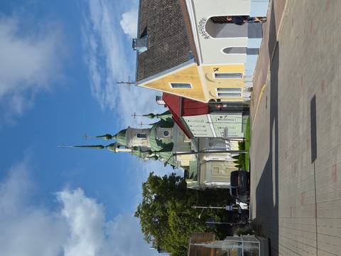       Quiet small-town street lined with pastel houses leading to a green-roofed orthodox church with tall spires.
  