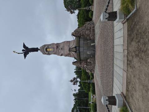       Angel statue with raised cross perched atop a rugged stone pedestal on a cloudy day.
  
