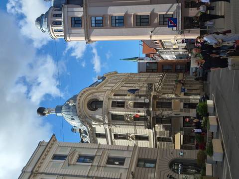       Art-nouveau corner building with ornate dome and clear blue sky, travelers with luggage walk the narrow Riga street.
  