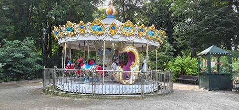       Colorful vintage carousel in a shady park clearing, surrounded by a metal fence and lush greenery.
  