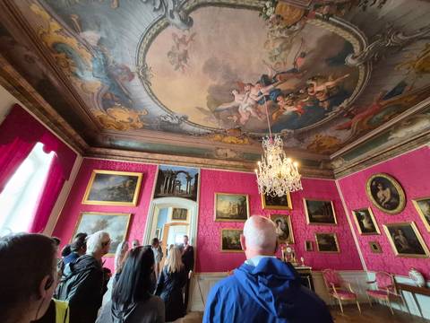       Lavish Baroque palace room with pink walls, gilded frames, and ceiling fresco visited by a tour group.
  