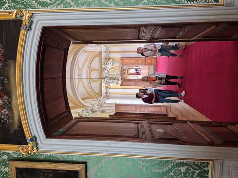       Visitors walking through an ornate palace corridor with arched ceilings and a rich red carpet.
  
