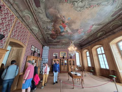       Elegant palace library with painted ceiling and chandelier where tourists observe exhibits and tile stove.
  
