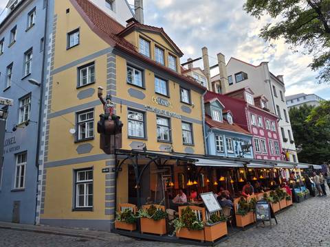       Colorful row of restaurants in Riga Old Town with outdoor diners beneath warm hanging lights.
  