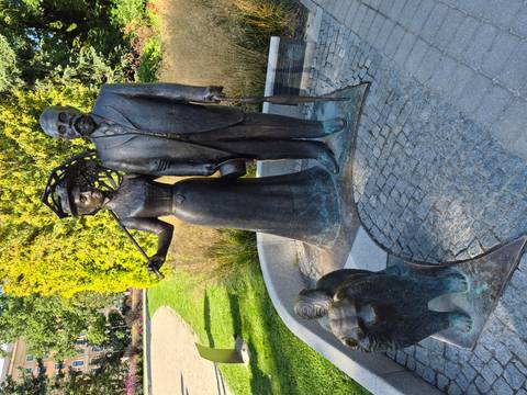       Bronze statue of an elegantly dressed couple with a dog in a sunny park setting.
  
