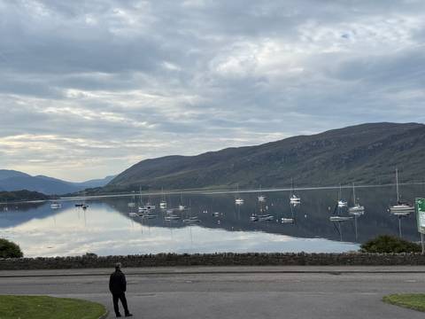       Still lake reflecting moored sailboats with surrounding mountains under a softly overcast sky.
  